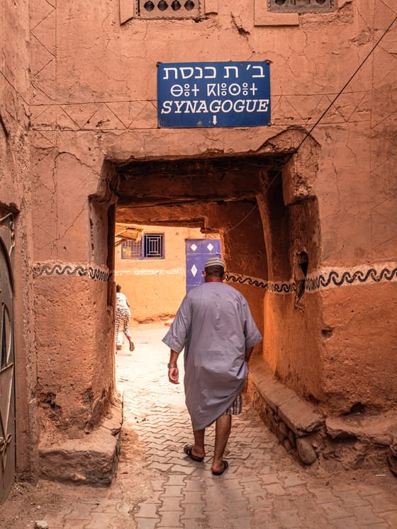 An earthen kasbah lane with a sign in Hebrew, Arabic and English marking a synagogue.