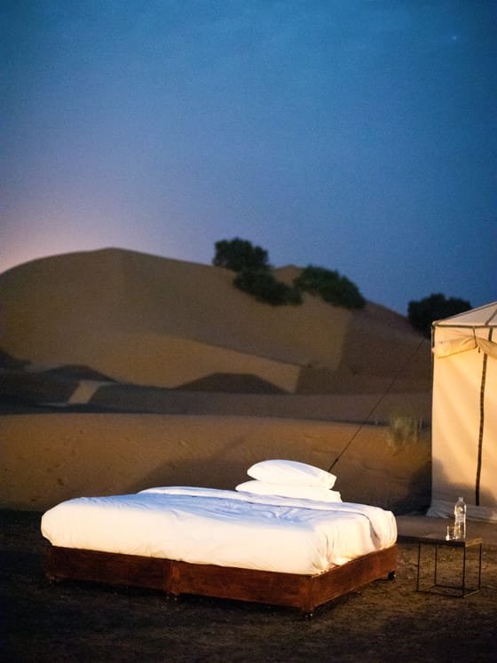 A bed made up under the open sky beside a desert tent at dusk.